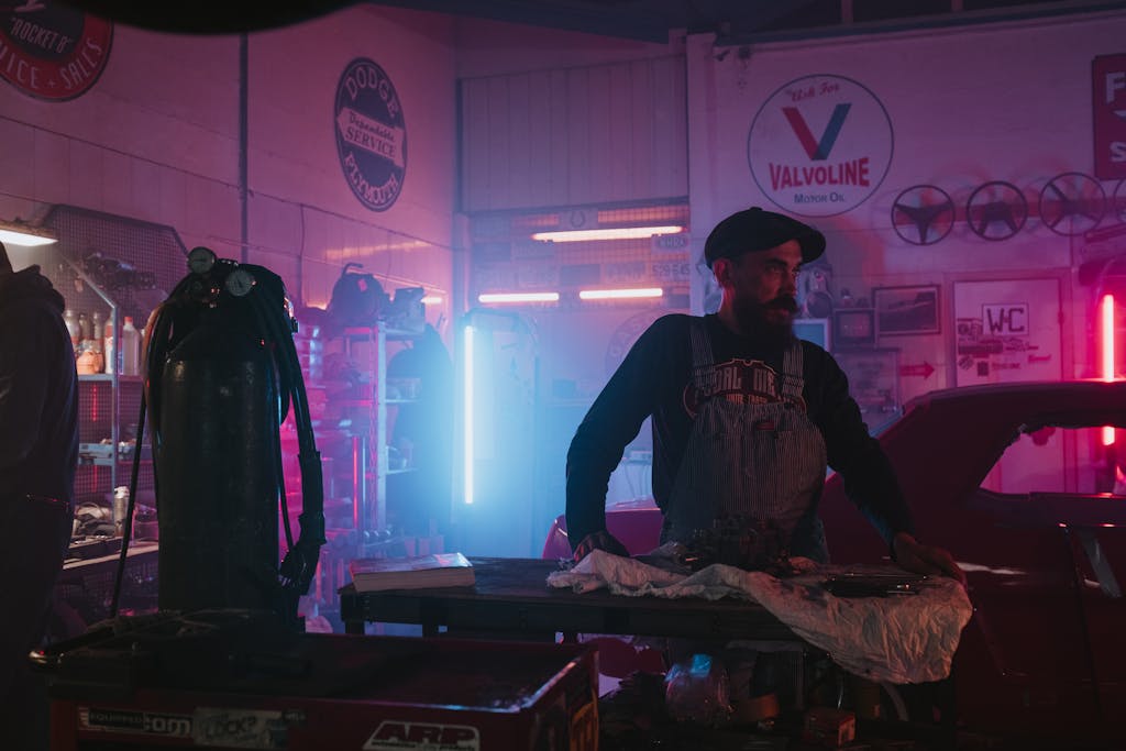 Mechanic works in a vintage garage with neon lighting and classic cars.